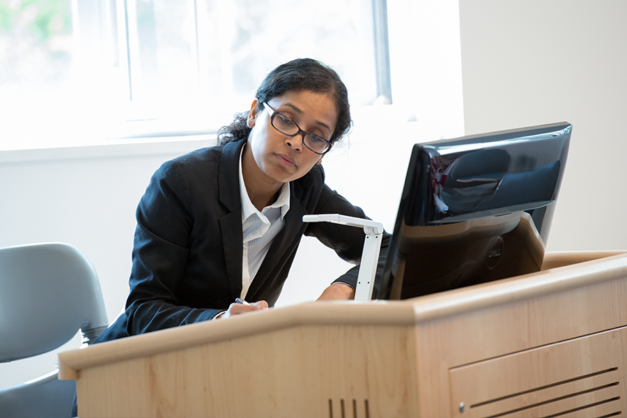 A faculty member stands at a lectern in front of a computer.