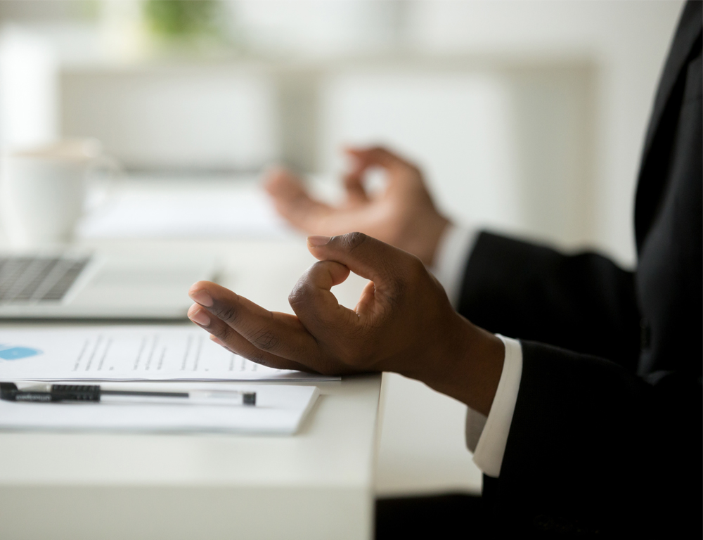 A closeup of a man’s forearms, which are resting on a desk where he is seated. The man is wearing a suit. On each hand, his index finger and thumb form a circle, with the other fingers outstretched.