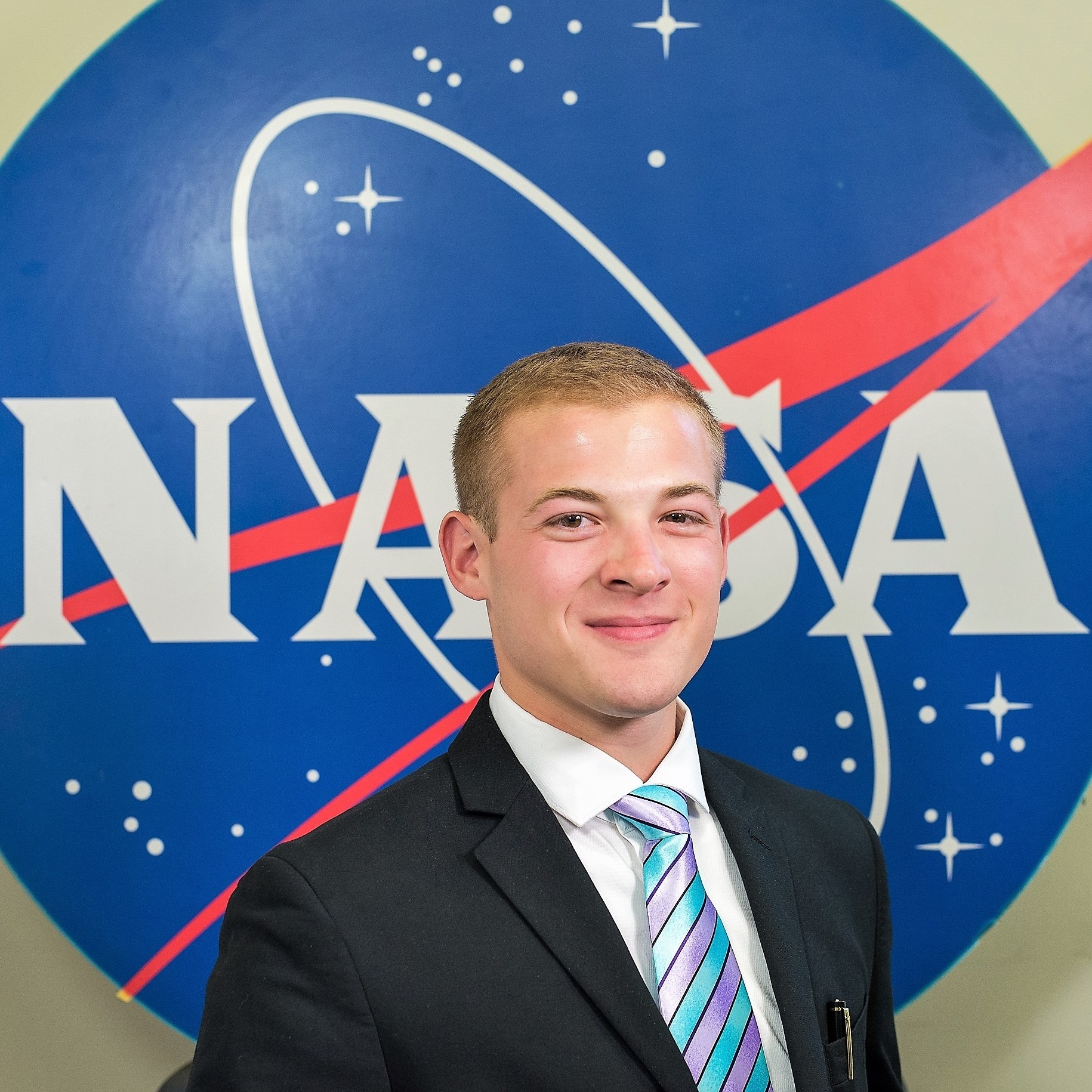 male student in business attire smiles and poses in front of the NASA logo. 