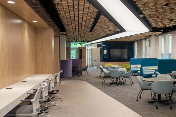 Photo of a room with a desk bar along the left wall, several booths and tables, a large monitor mounted on a blue glass wall, and an entryway at the end of the room.