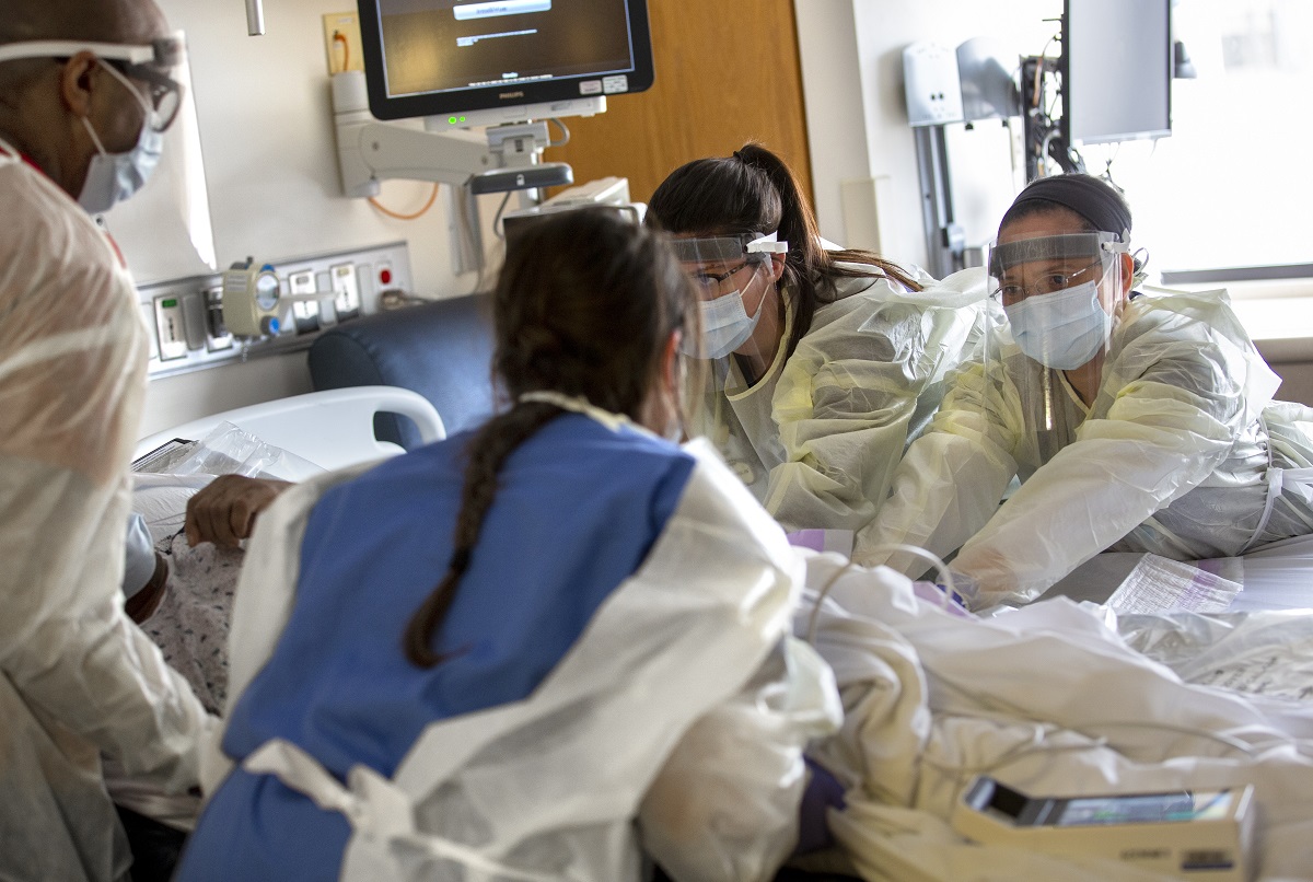Two nurses, facing camera, with the Medical Intermediate Care Unit at Hershey Medical Center, and two other health care workers, whose backs are to the camera, touch a patient who is lying in bed. They are wearing gowns and face shields.