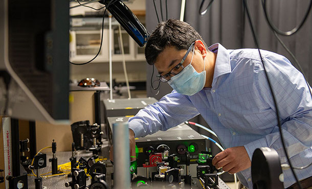 A man in a blue button-down shirt and a blue face mask adjusts optical devices that are shining green lights 