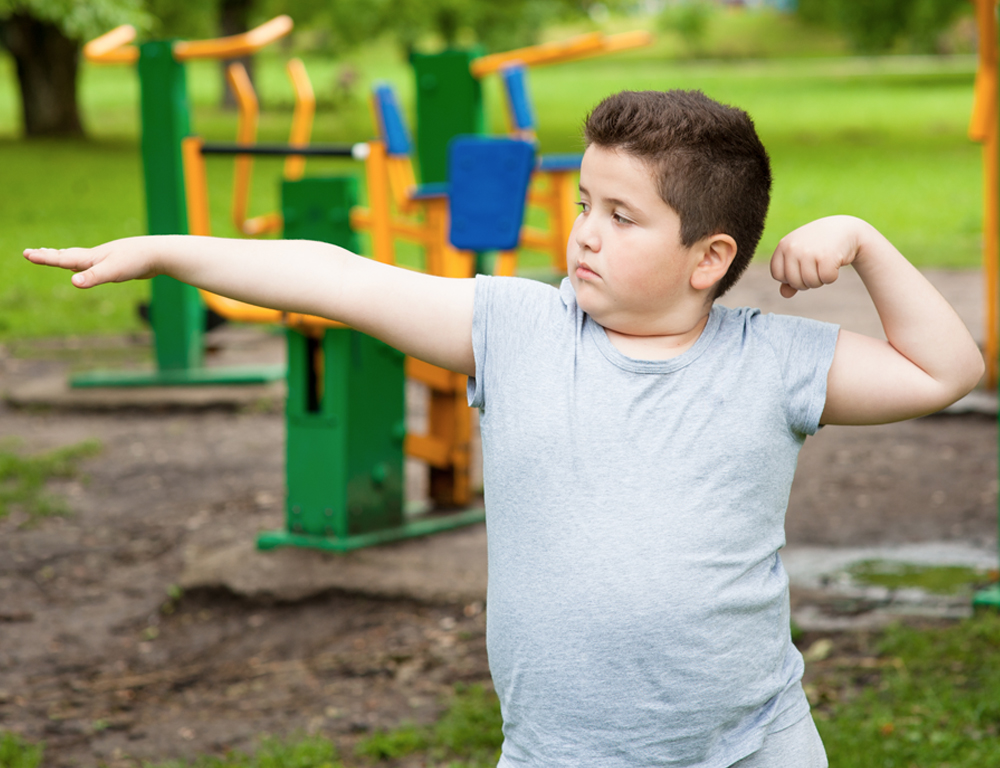 A young boy stands with his right arm extended outward and his left arm curled upward. Playground equipment is in the background.