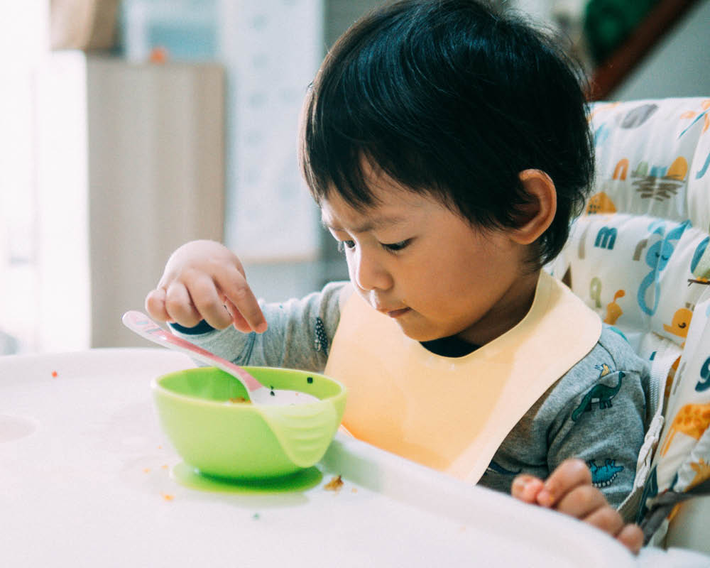 A young child sits at a table, a bowl and spoon in front of them