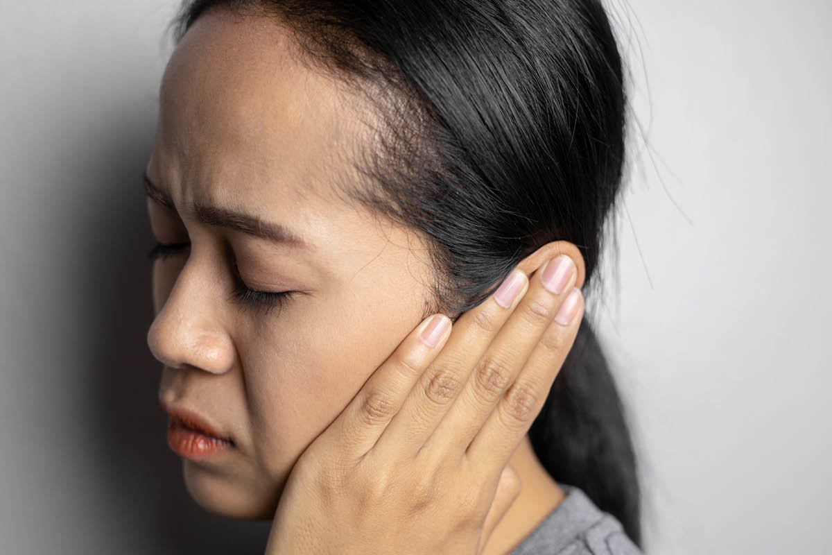 A close-up of a woman with her eyes closed and an irritated expression on her face. She is covering her left ear with her left hand.