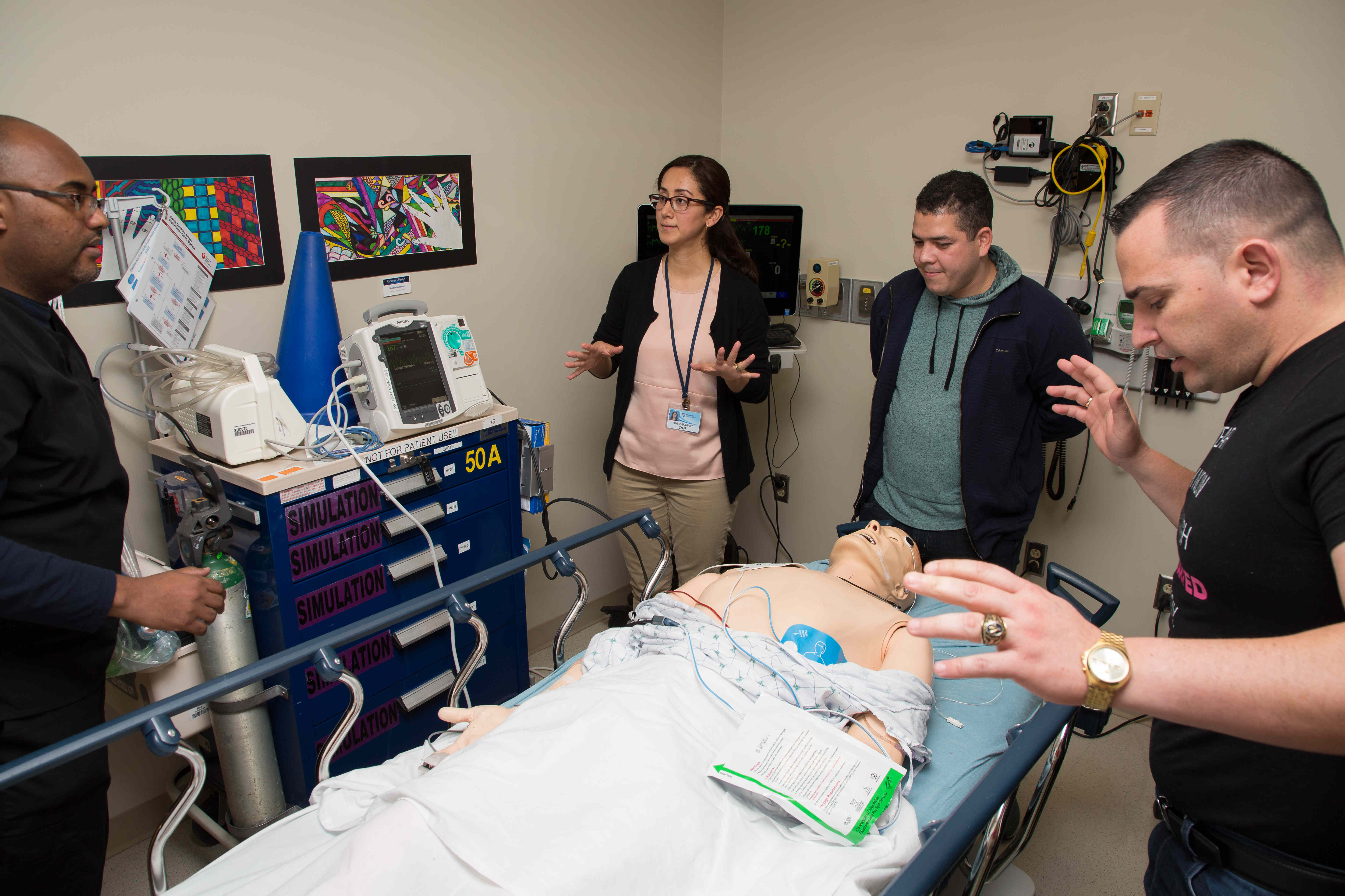 Dr. Lidys Rivera Galvis and students stand around a mannequin that lays on a wheeled-stretcher and is connected to heart machine.