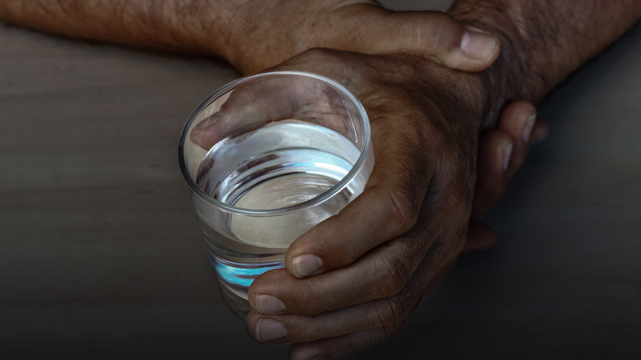 Two hands on a table, the right hand holds the left wrist, and the left hand holds a glass of water.  