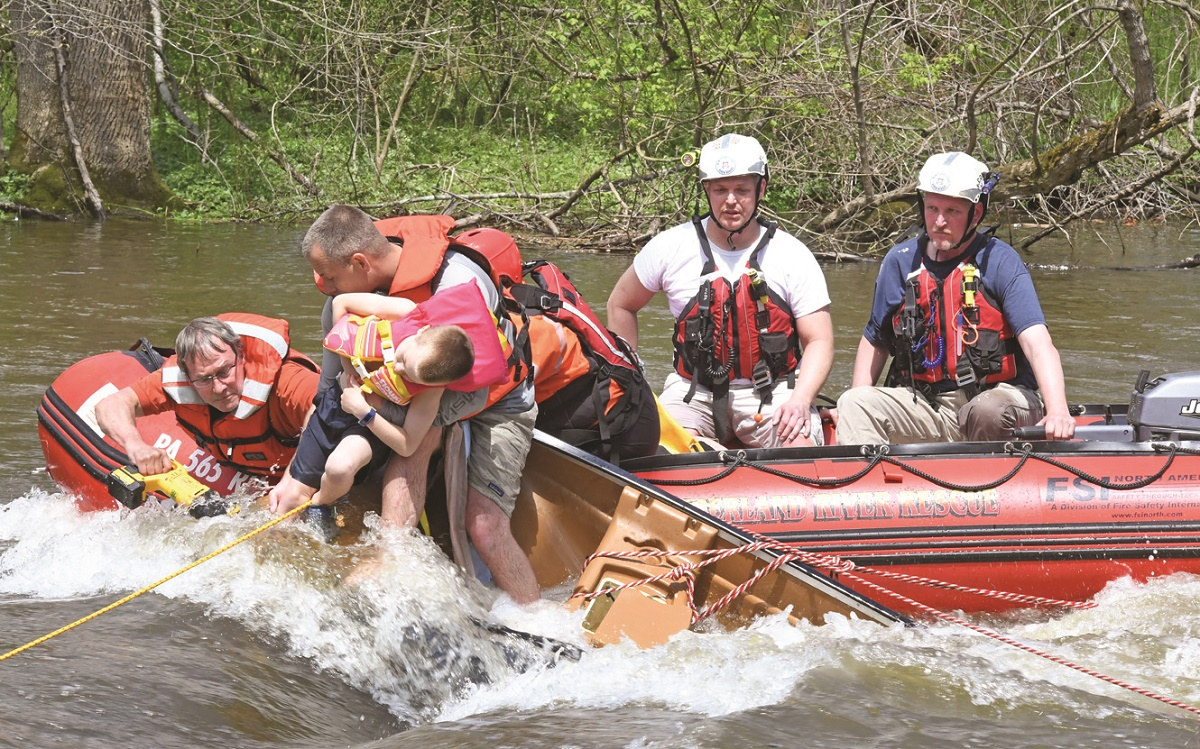 Rex Carmichael, wearing glasses and a life vest, saws tree roots to free a child in a creek as another paramedic holds onto the boy, who is slumped over his arm. Three other paramedics, wearing helmets and life jackets, sit in a river rescue boat.
