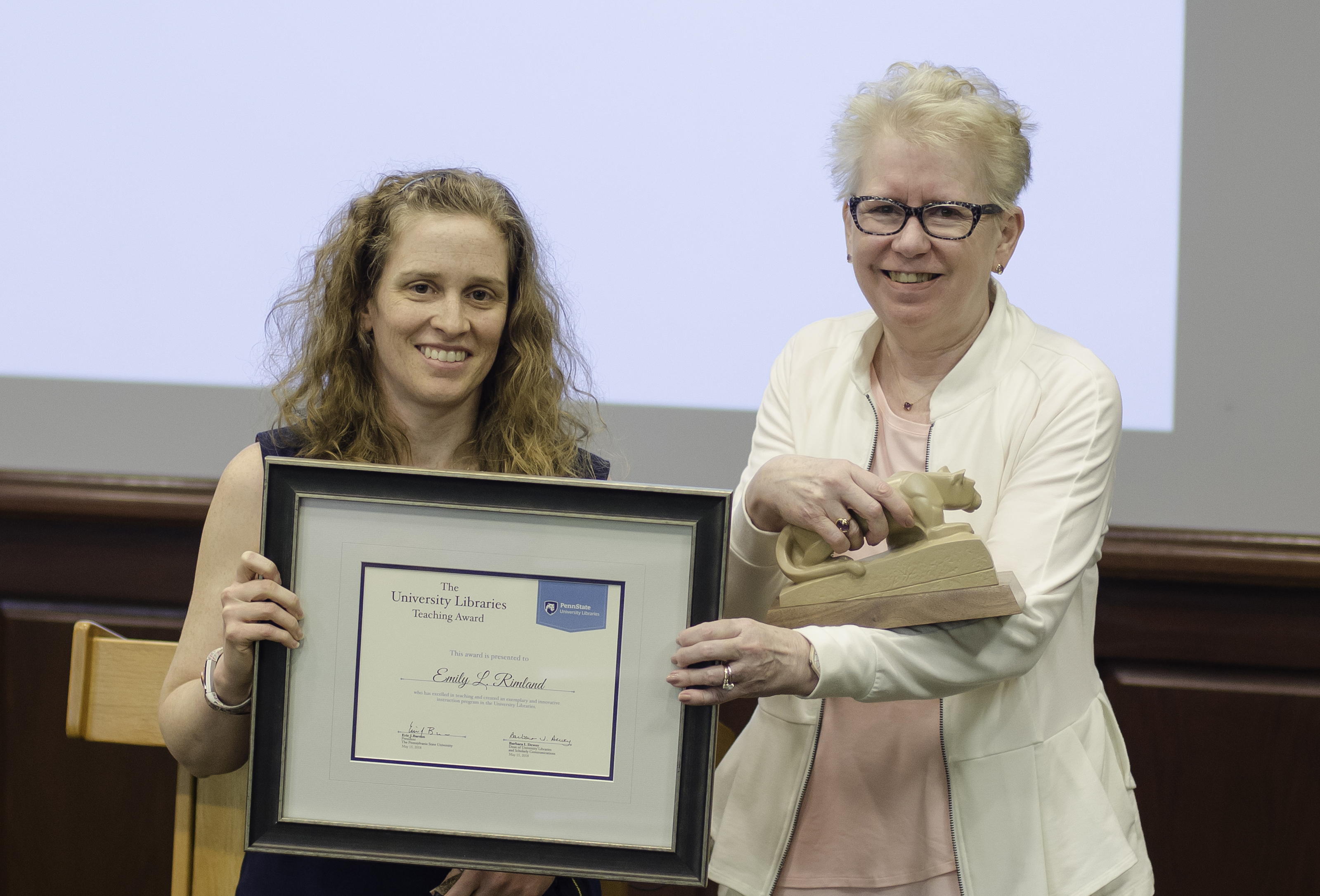 two women standing, one holding award certificate, other presenting small sculpture of Nittany Lion as award