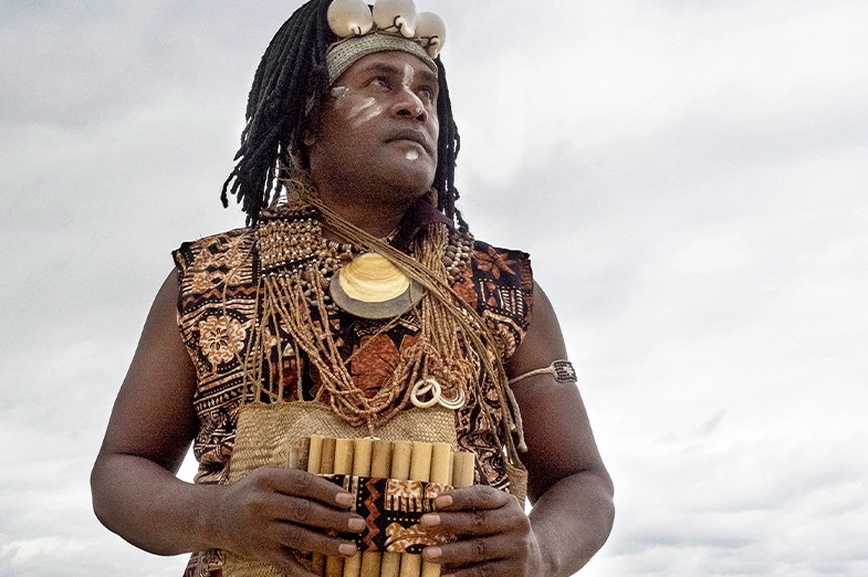 A man wearing shells and beads on his bare chest holds a pan pipe.