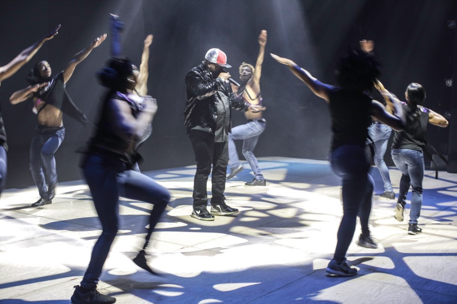 A group of dancers move in a circle around a man singing into a microphone.