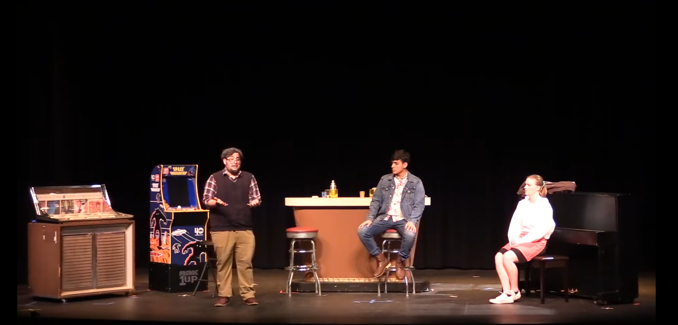 two men are sitting at a bar, one man talking animatedly, and a woman is sitting backward at a piano bench listening