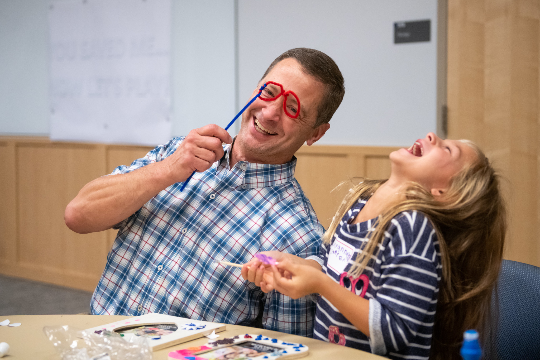 Dr. Christopher O’Hara holds up pipe cleaners twisted into two circles on a straight length of pipe cleaner and leans toward Savannah Doster, who throws back her head and laughs. The two are seated at a table upon which are frames and craft materials.