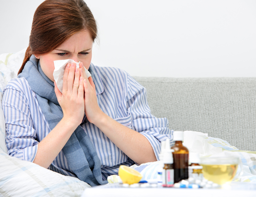 A woman blows her nose with a tissue. In the foreground is a table with various liquid medications and pills, as well as a drink and other items.