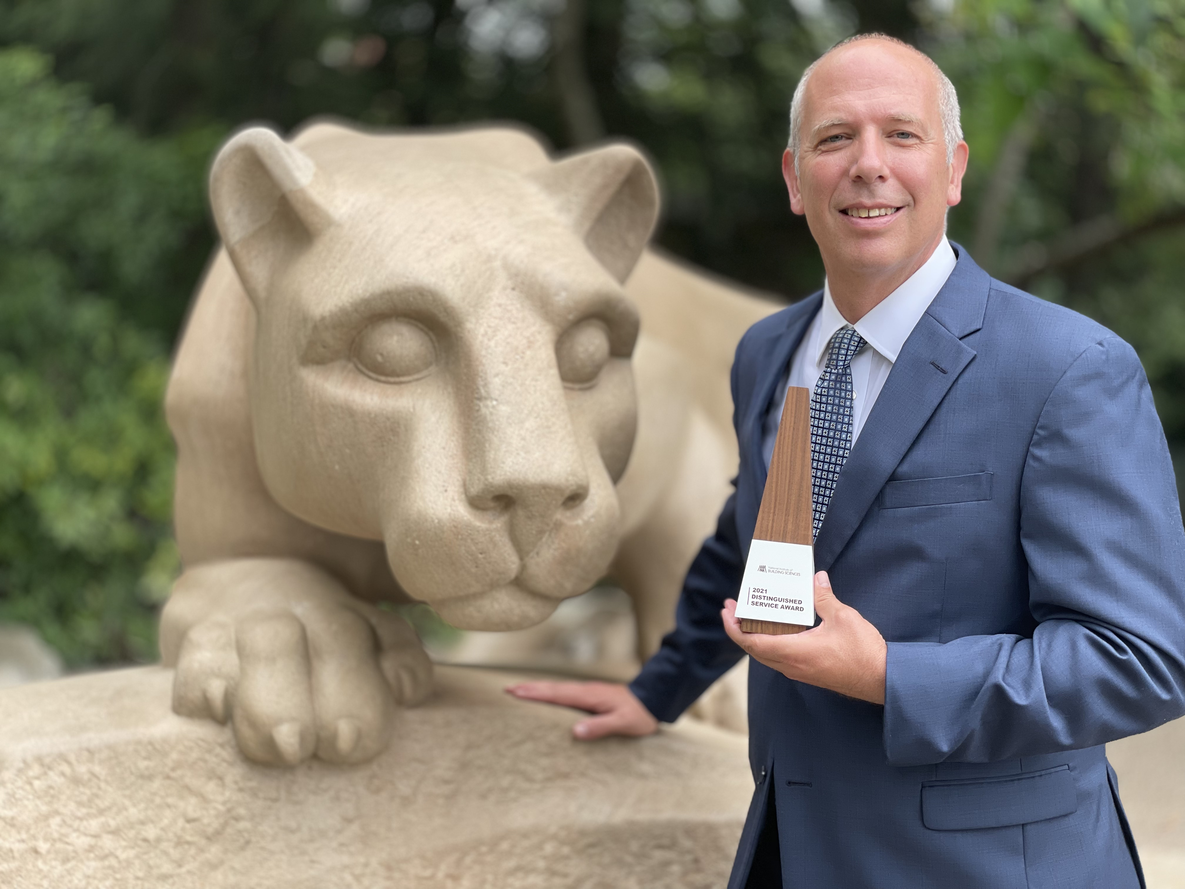 A man in a tie smiles at the camera and holds a brown triangular award. The Nittany Lion statue is visible behind him. 