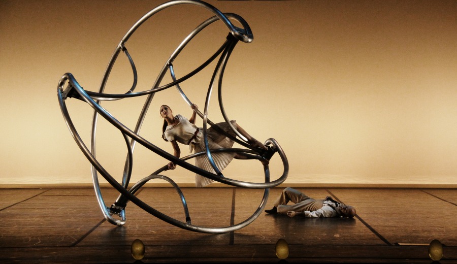 An acrobat is positioned on the Cyr Wheel while another lies on the ground nearby.