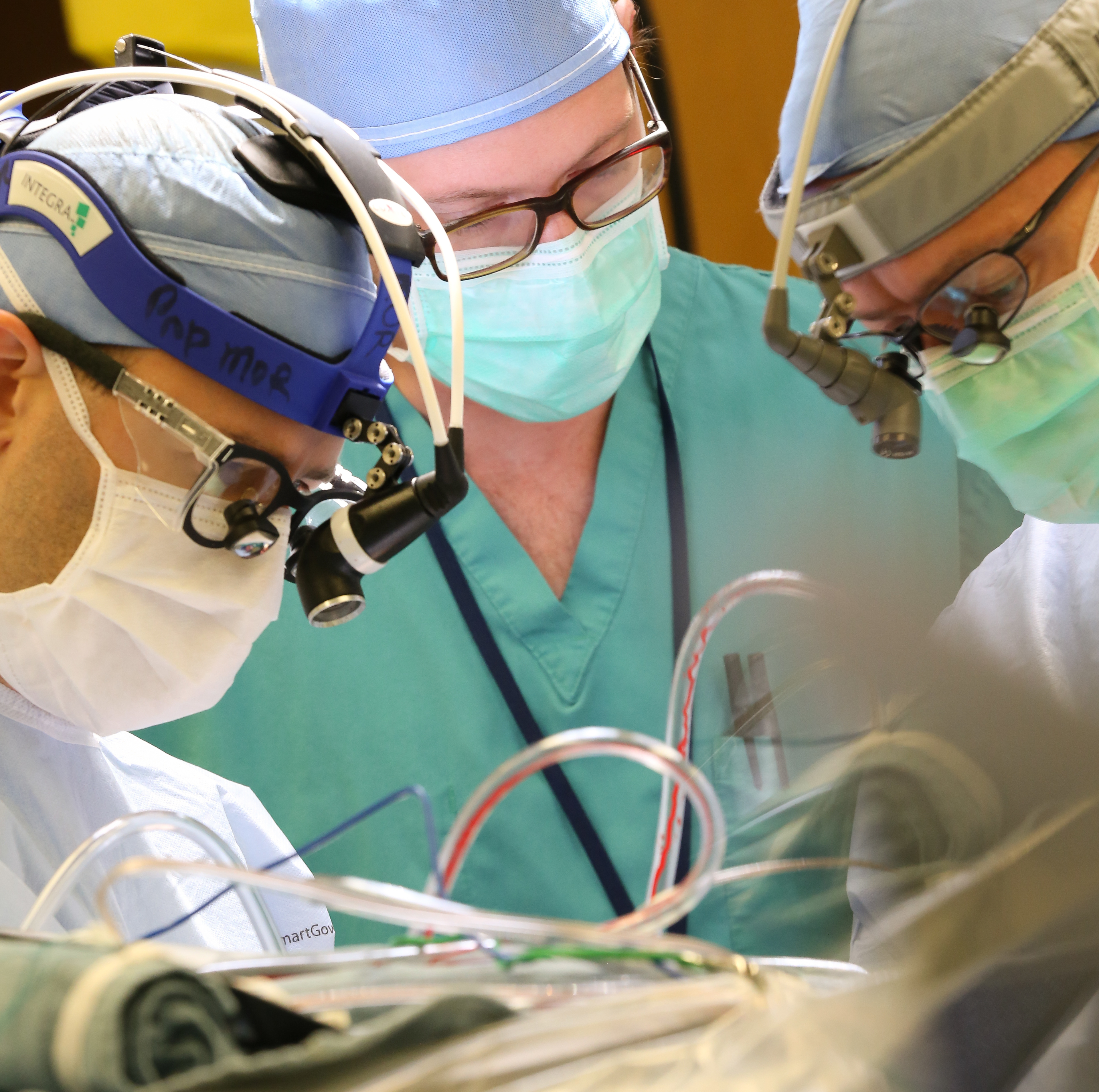 Three surgeons/medical staff work in the operating room, over a patient. The patient is not visible, but tubes emerge out of the bottom of the photo.