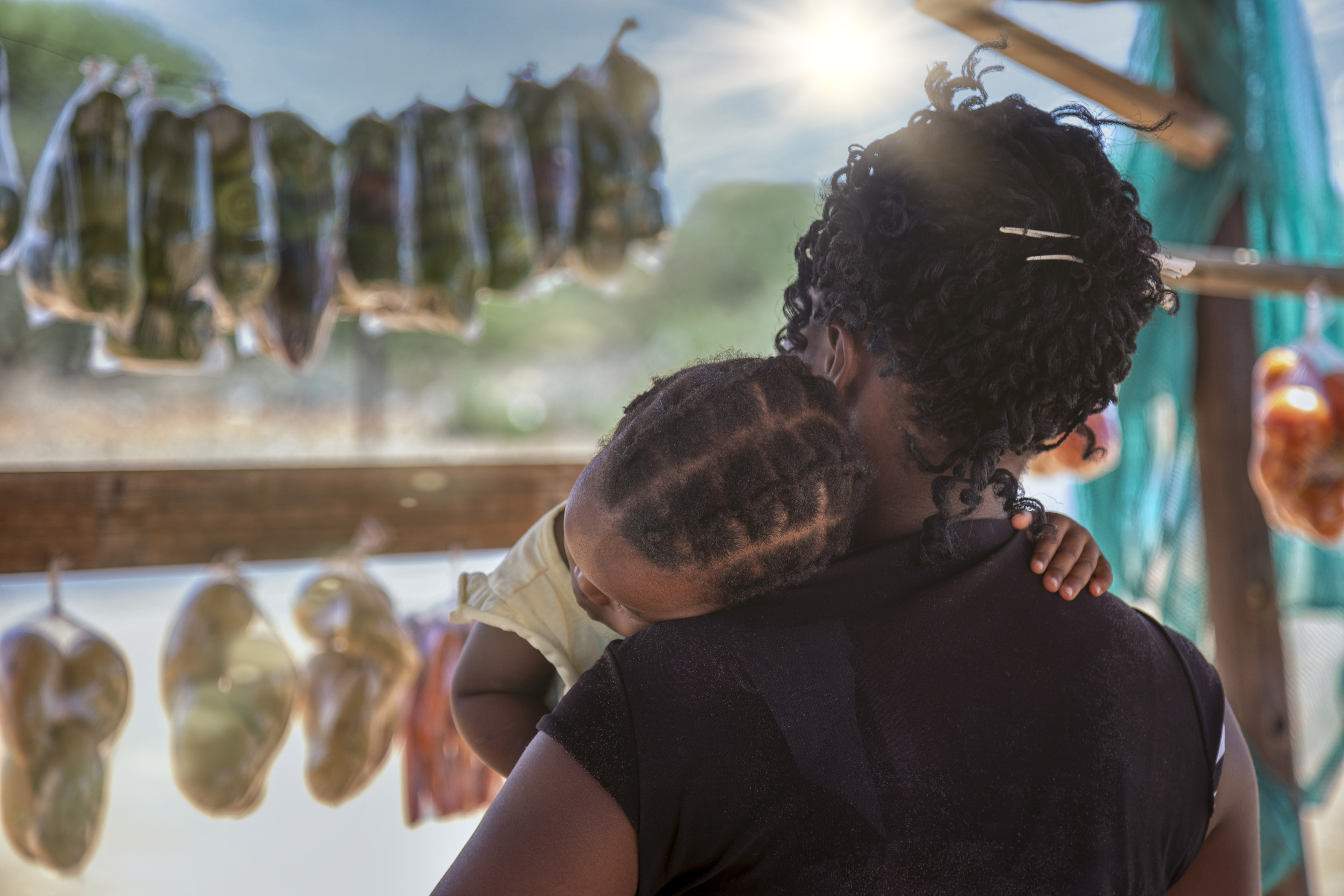 Woman seen from behind holding small child in her arms as she browses for food in an African market