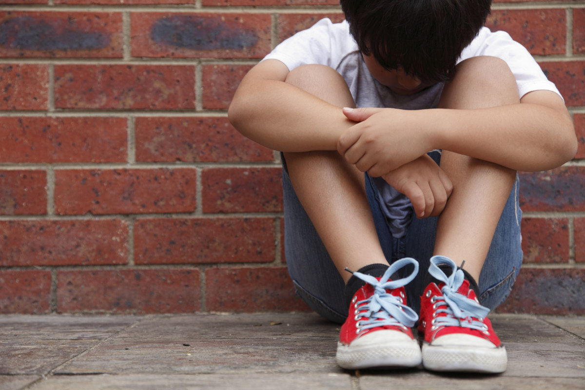 A young boy sits on a sidewalk, leaning against a brick wall, looking down at the ground.