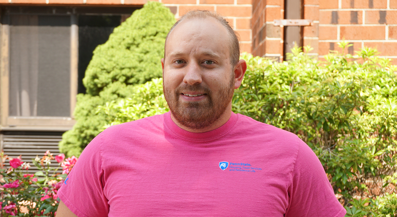 Smiling man in a pink t-shirt standing outside a brick building with green bushes behind him.