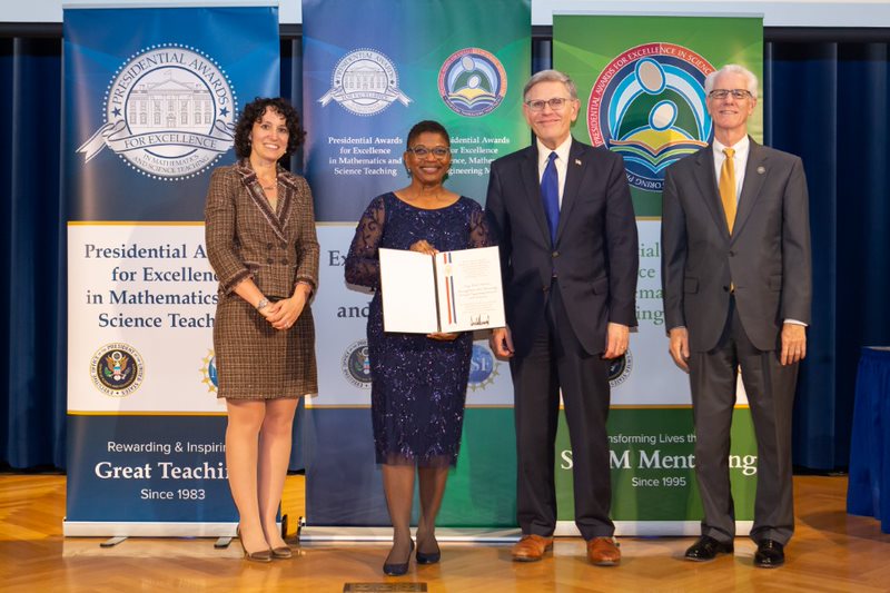 Two women and men stand on a stage, smiling at the camera. The African-American woman holds an award. 