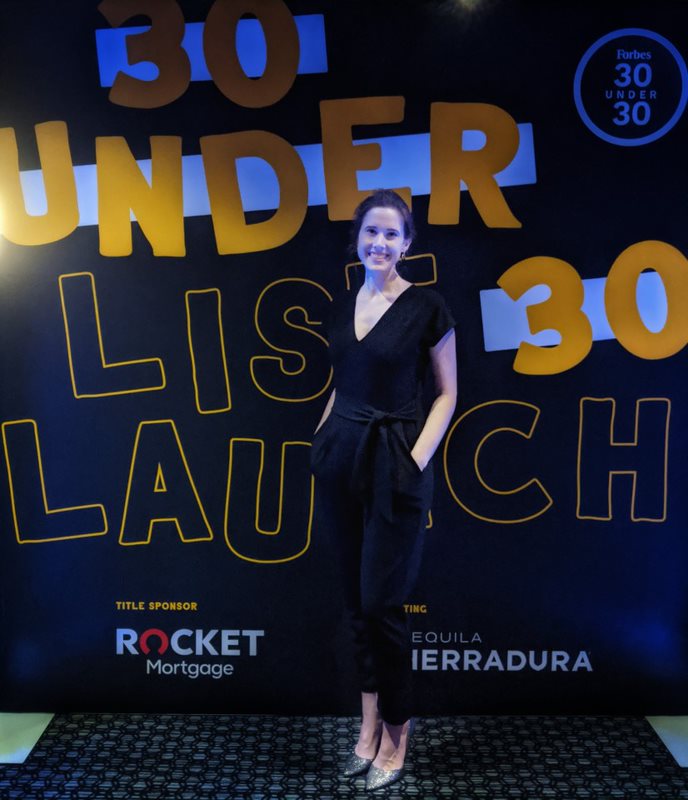 A Caucasian woman poses in front of signage that reads "Forbes 30 Under 30 Launch."