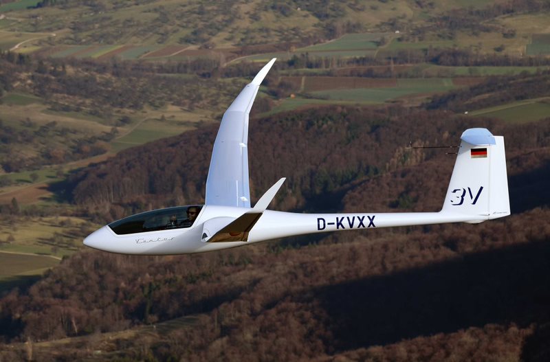 A smiling man gives a thumbs up from the cockpit of a small glider plane flying over forest. 