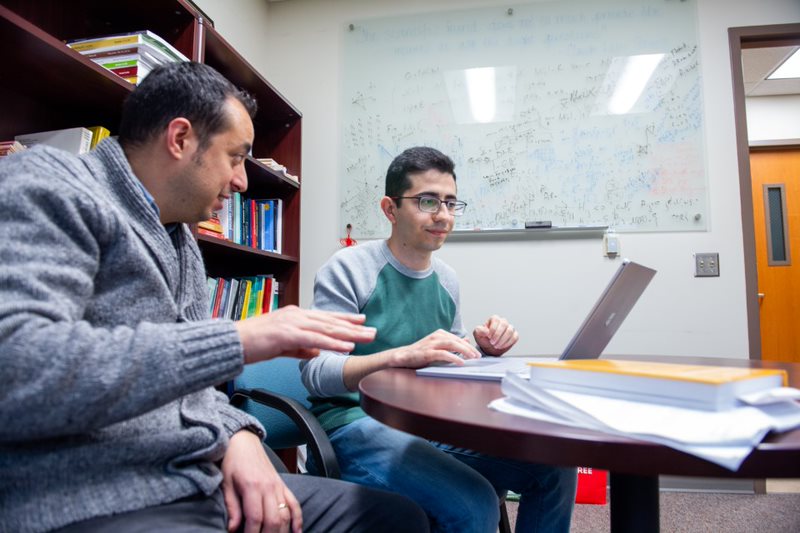 Man in a gray sweater speaks to man in green shirt while looking at laptop.