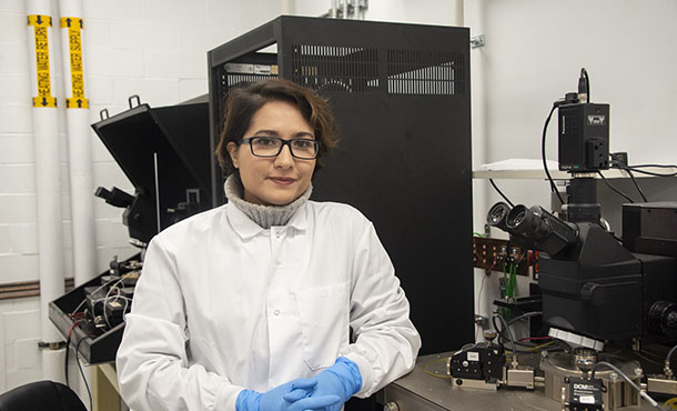 A woman in a white lab coat, blue gloves, and glasses sits next to a microscope in a lab and smiles at the camera 