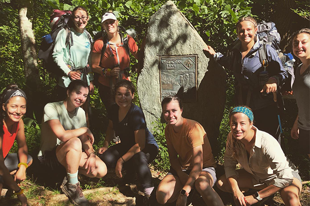 Nine women dressed in hiking gear pose for a photo near a rock. 