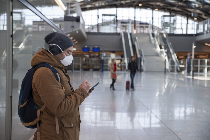 A man wearing a face mask stands alone in a railway station while using his smartphone