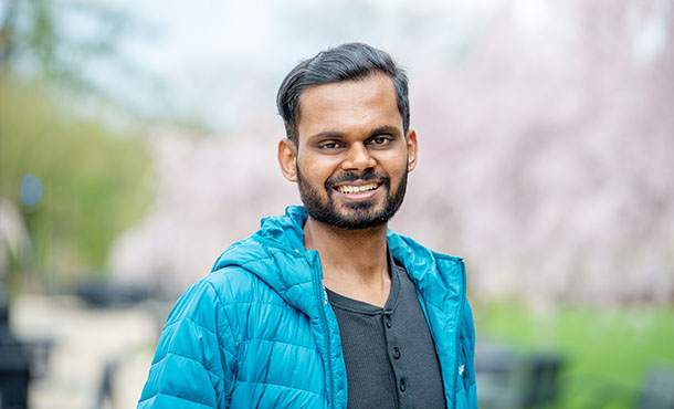 A man wears a light blue jacket and smiles at the camera in front of a blurred background of cherry blossom trees 