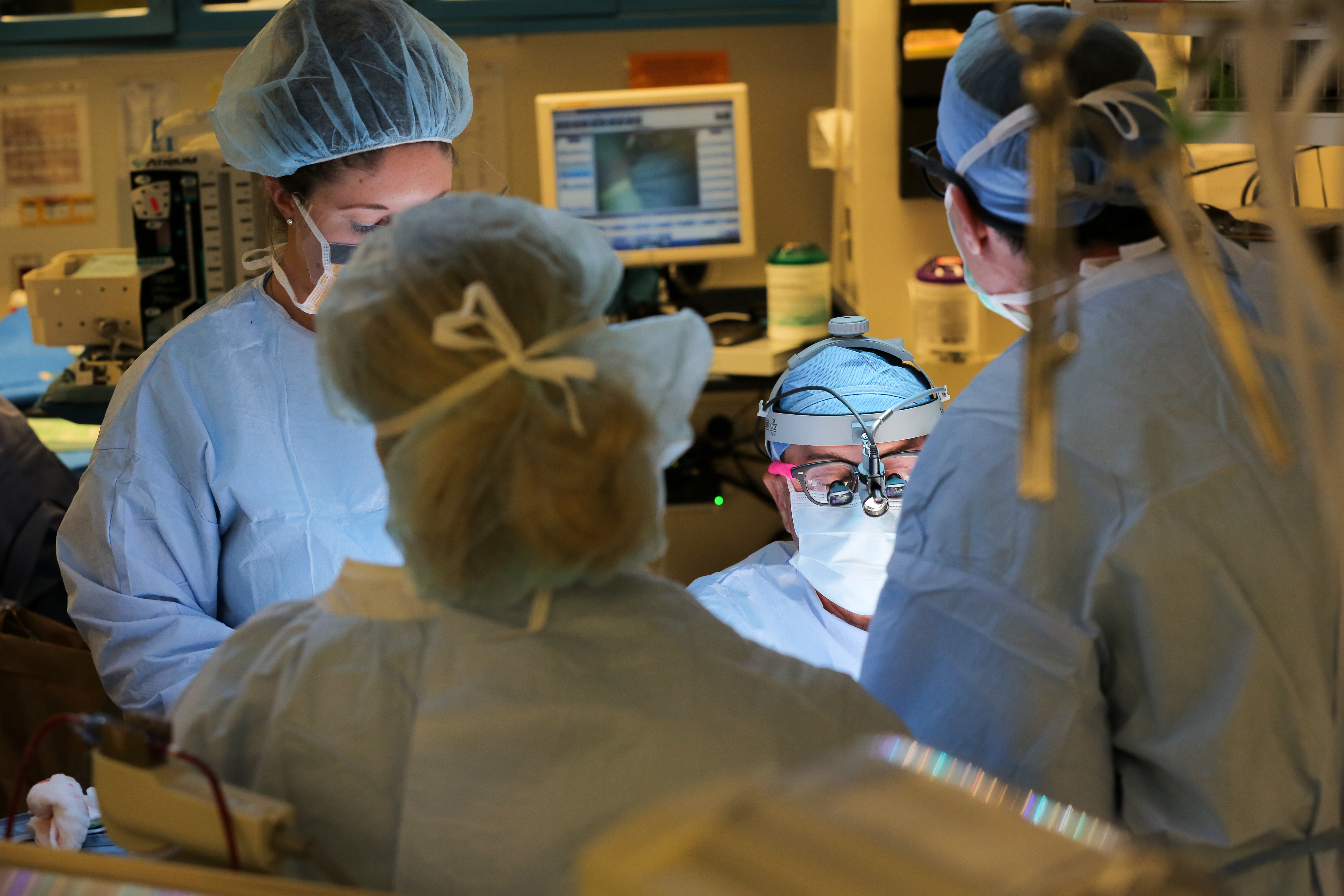 Four people in surgical scrubs and gowns are gathered around a patient in the operating room. A computer monitor and some equipment are visible in the background.