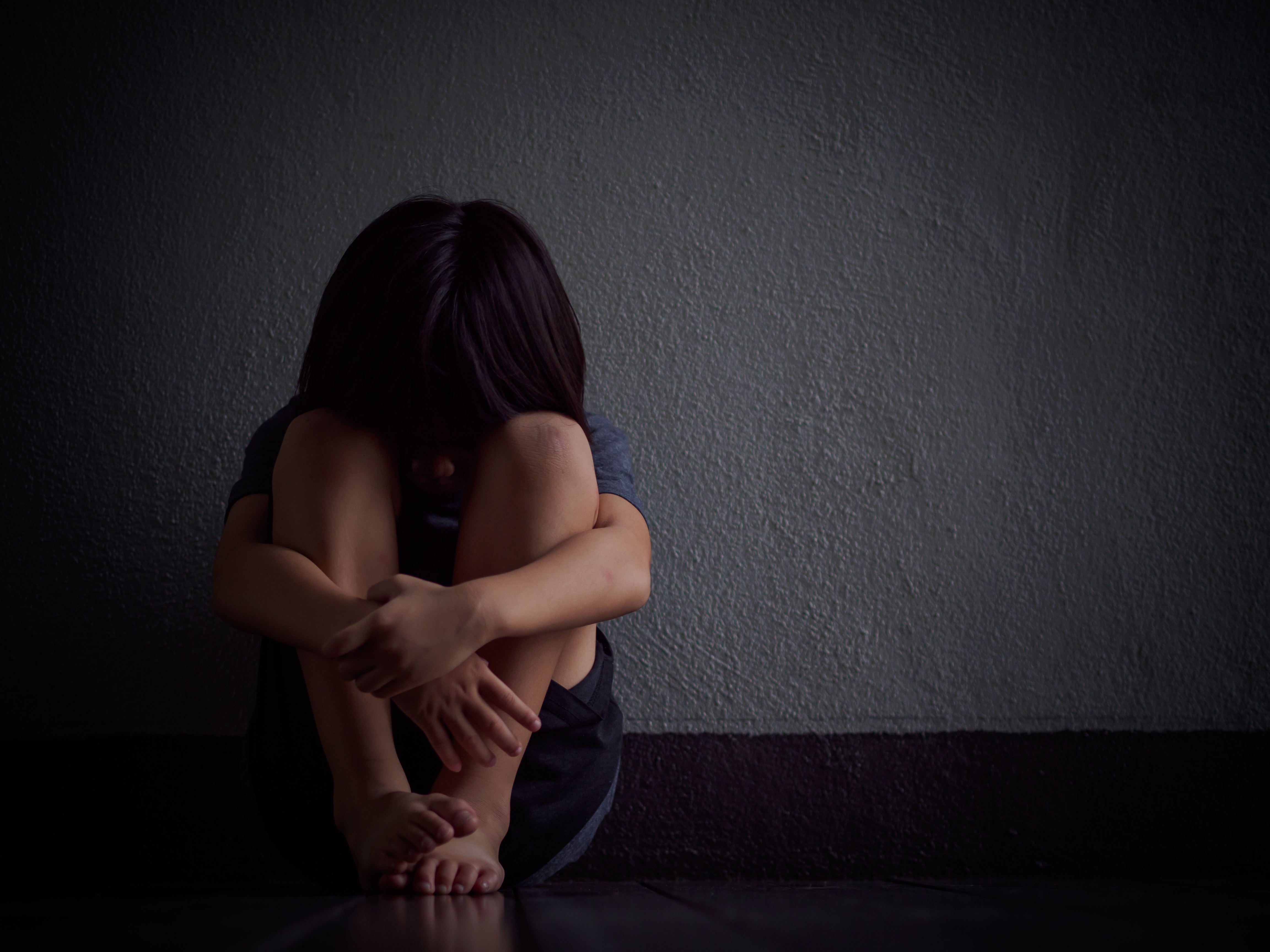 A little boy unhappy and alone sitting against a wall in a dark room with his head against his knees