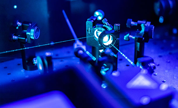 Beams of blue light shine out from a laser made of two optical lenses. Two similar lenses are on either side of the laser, and the entire image is tinted blue. 