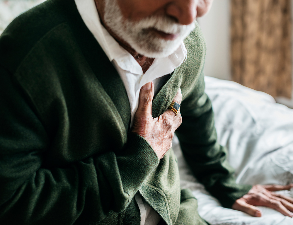 A close-up of a seated man, leaning slightly forward and clutching at his chest with his right hand.