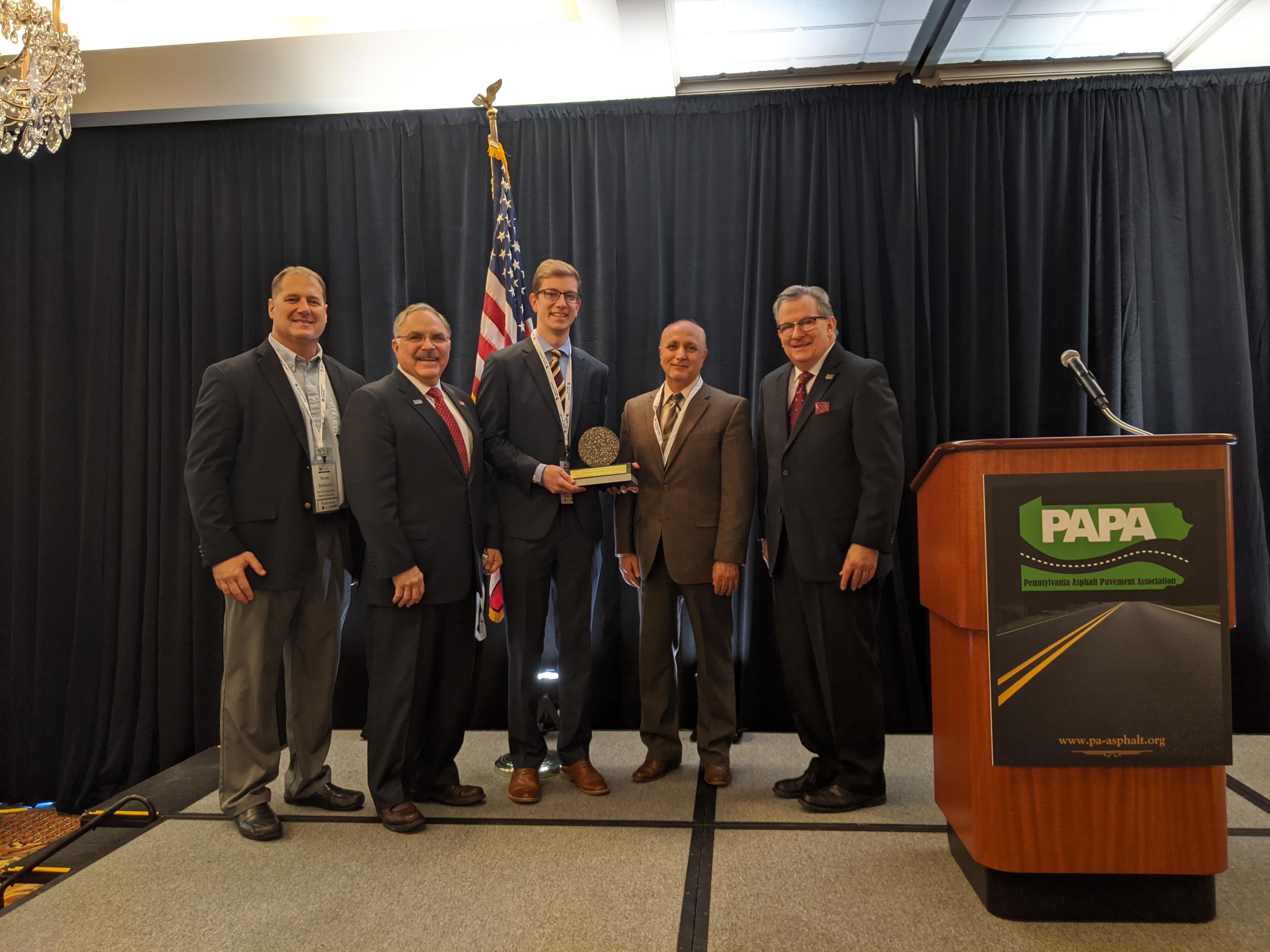 Five men dressed in business suits standing on a stage next to a podium. The man in the center is holding a trophy.