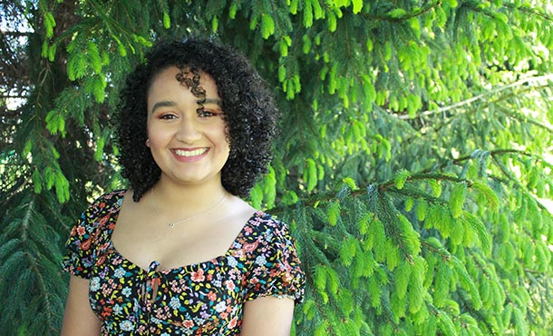 A young woman wearing a floral and paisley print top smiles at the camera in front of a green tree. 