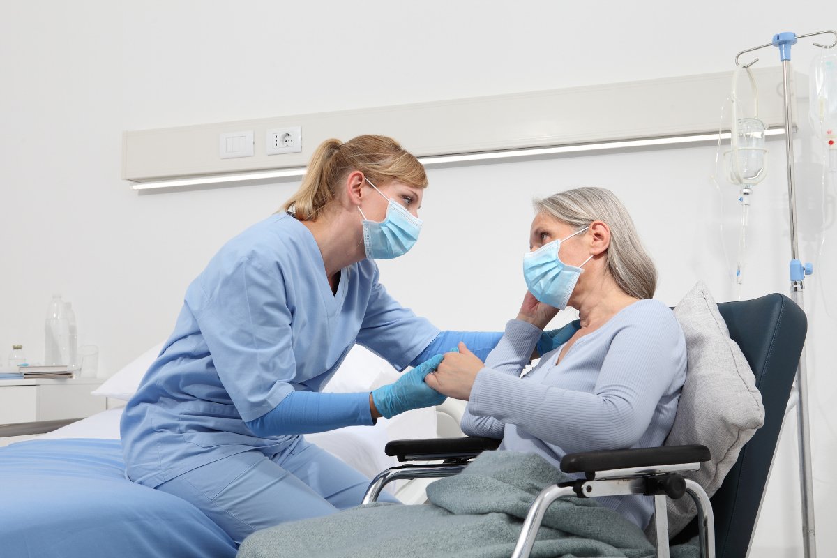 A nurse comforts an elderly woman in a wheelchair by holding her hand and placing her other hand on the patient’s shoulder.