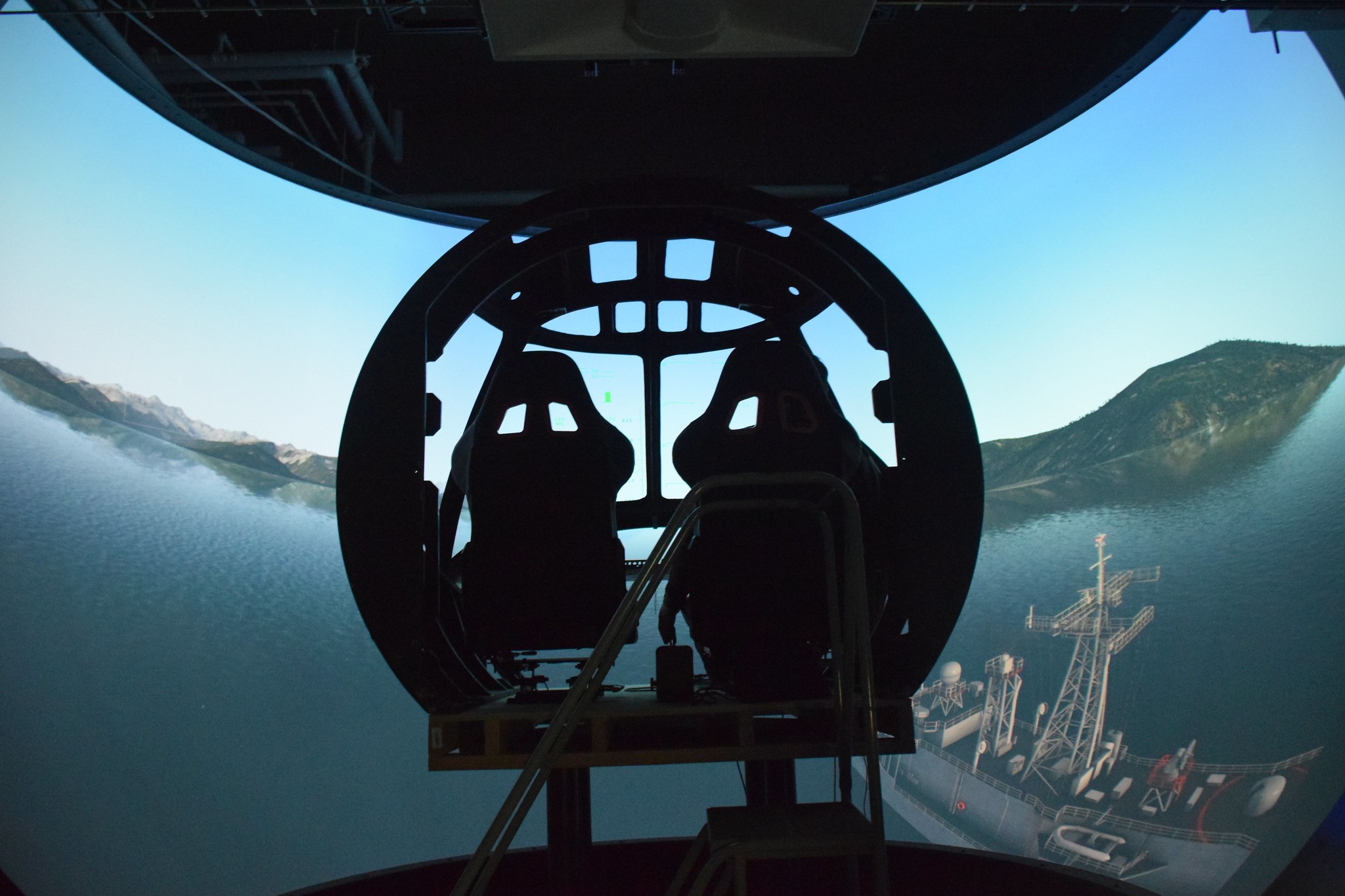 A cockpit is shadowed against a simulated water scene over a ship with mountains in the background. 