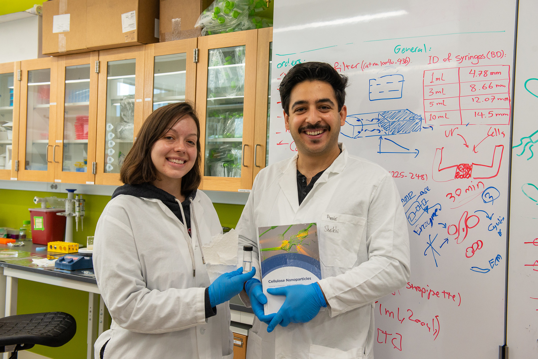 A man and woman in lab coats smile in front of a white board. The woman is holding paper, while the man holds a book.