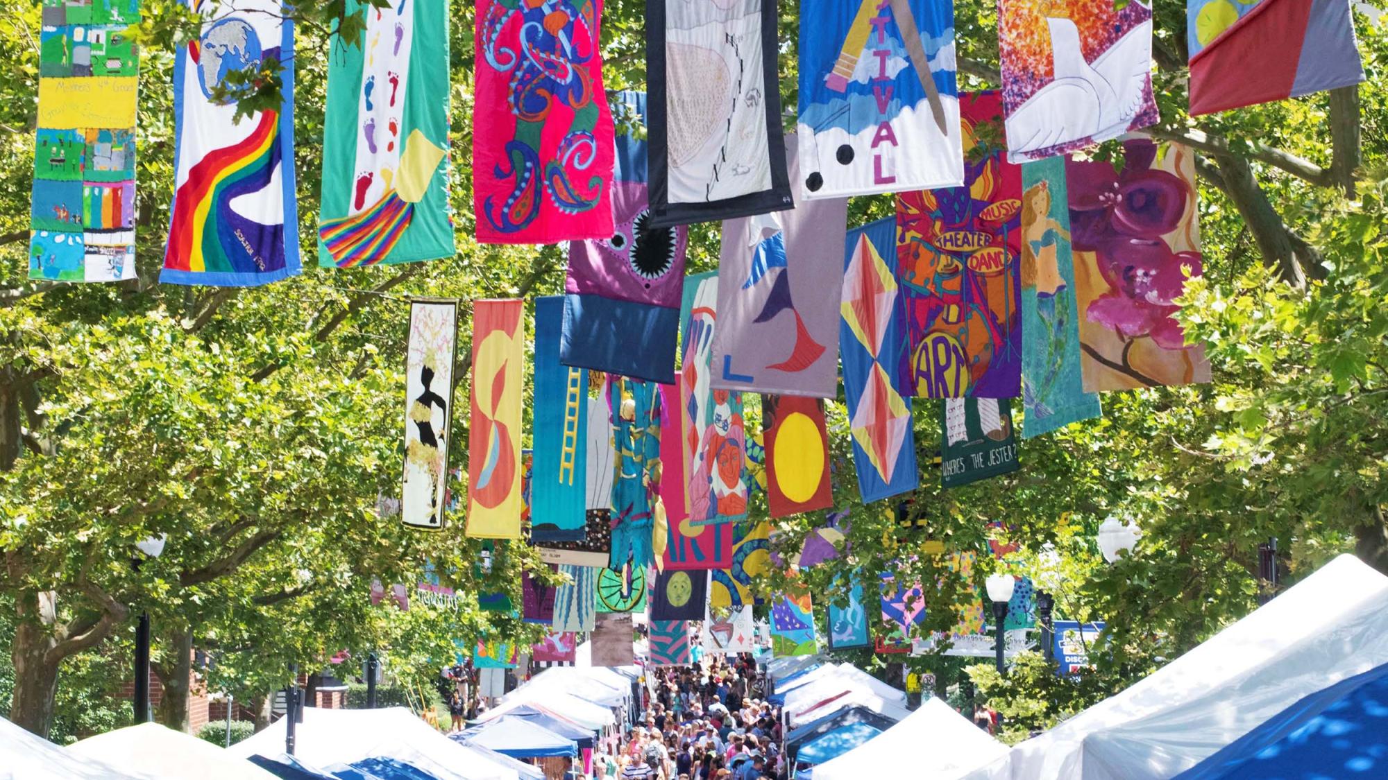 A crowd mills about on a tree-lined street, with colorful flags hanging overhead.