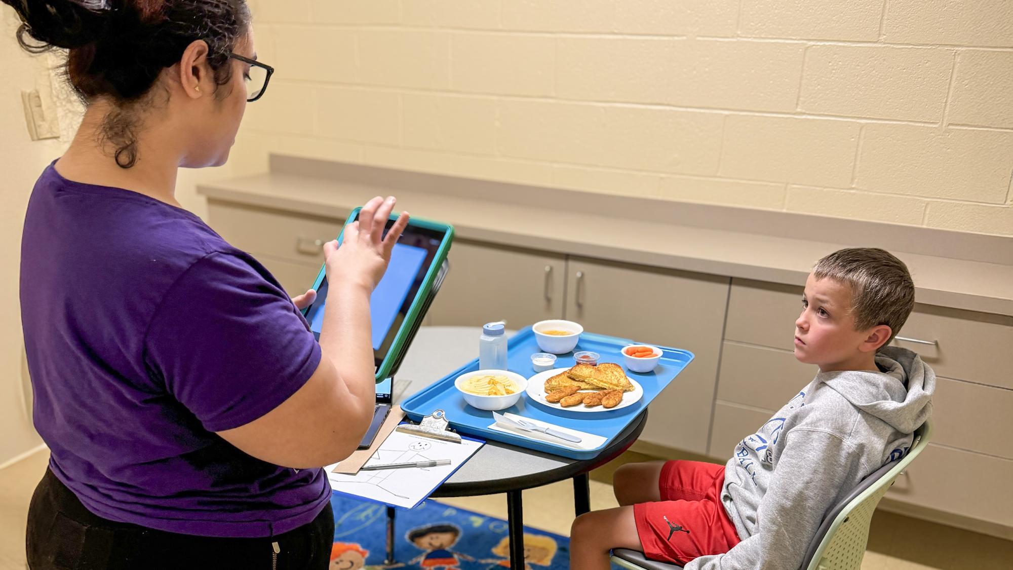 A child sits at a table with one of the sample meals used in the study and looks at a research assistant prior to eating.