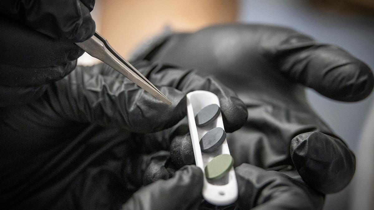 Gloved hands hold tweezers and a silver tray of three small circular ceramic tiles.