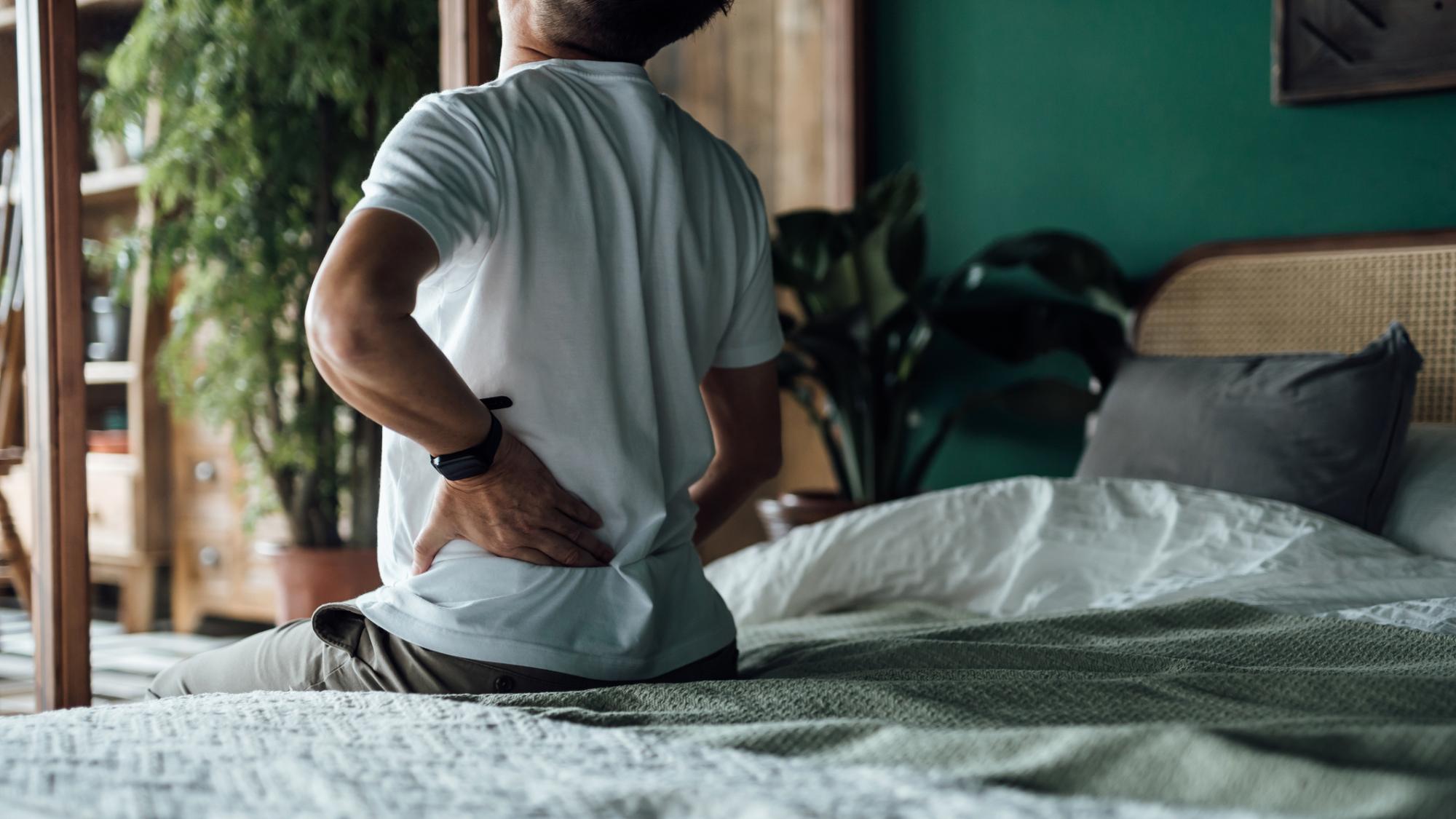 Older person sits on the edge of a bed with a hand on their back, indicating discomfort