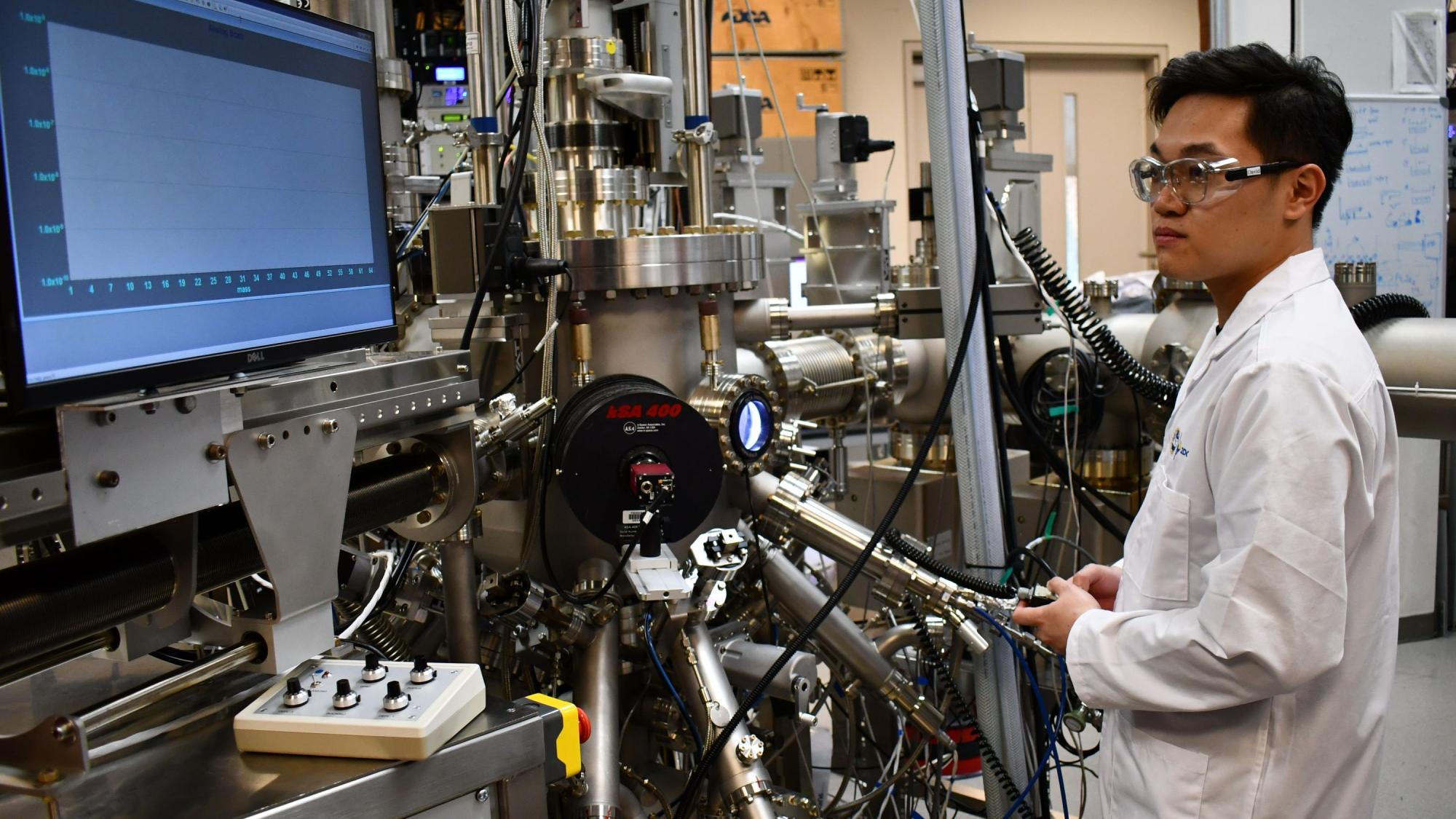 Person in lab coat and glasses operating a lab machine and looking at a monitor