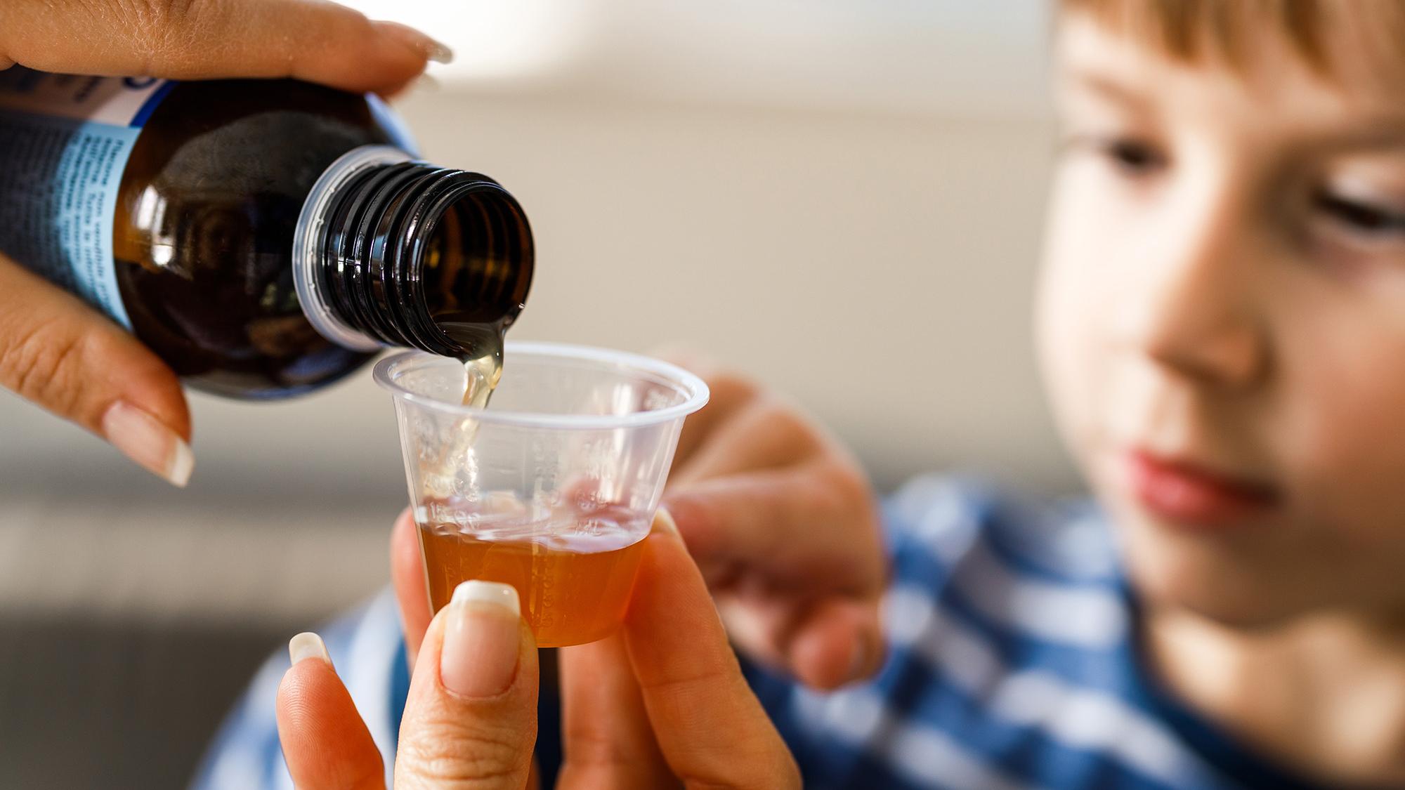Hands pouring liquid medication into a small cup in the foreground. A child is visible in the background.