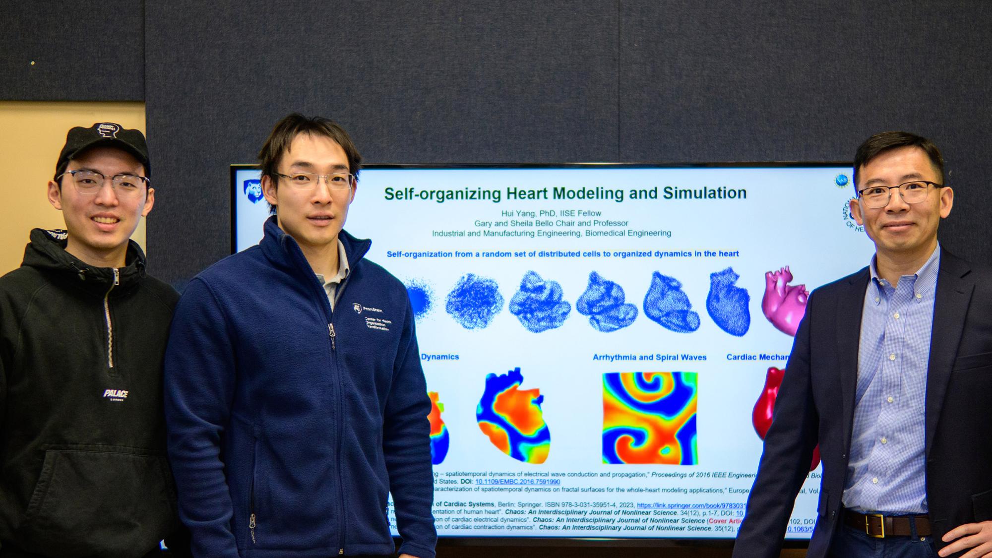 Three men pose for a photo in front of a television that reads "Self-organizing Heart Modeling and Simulation" with a series of graphics of graphics showing the heart in different phases of self-organization, with electrical maps of a heart beneath it. 