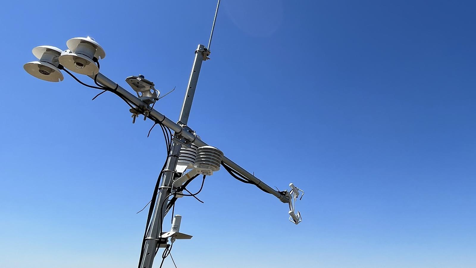A cross-shaped piece of equipment with attached sensors next to a field of crops against the blue sky