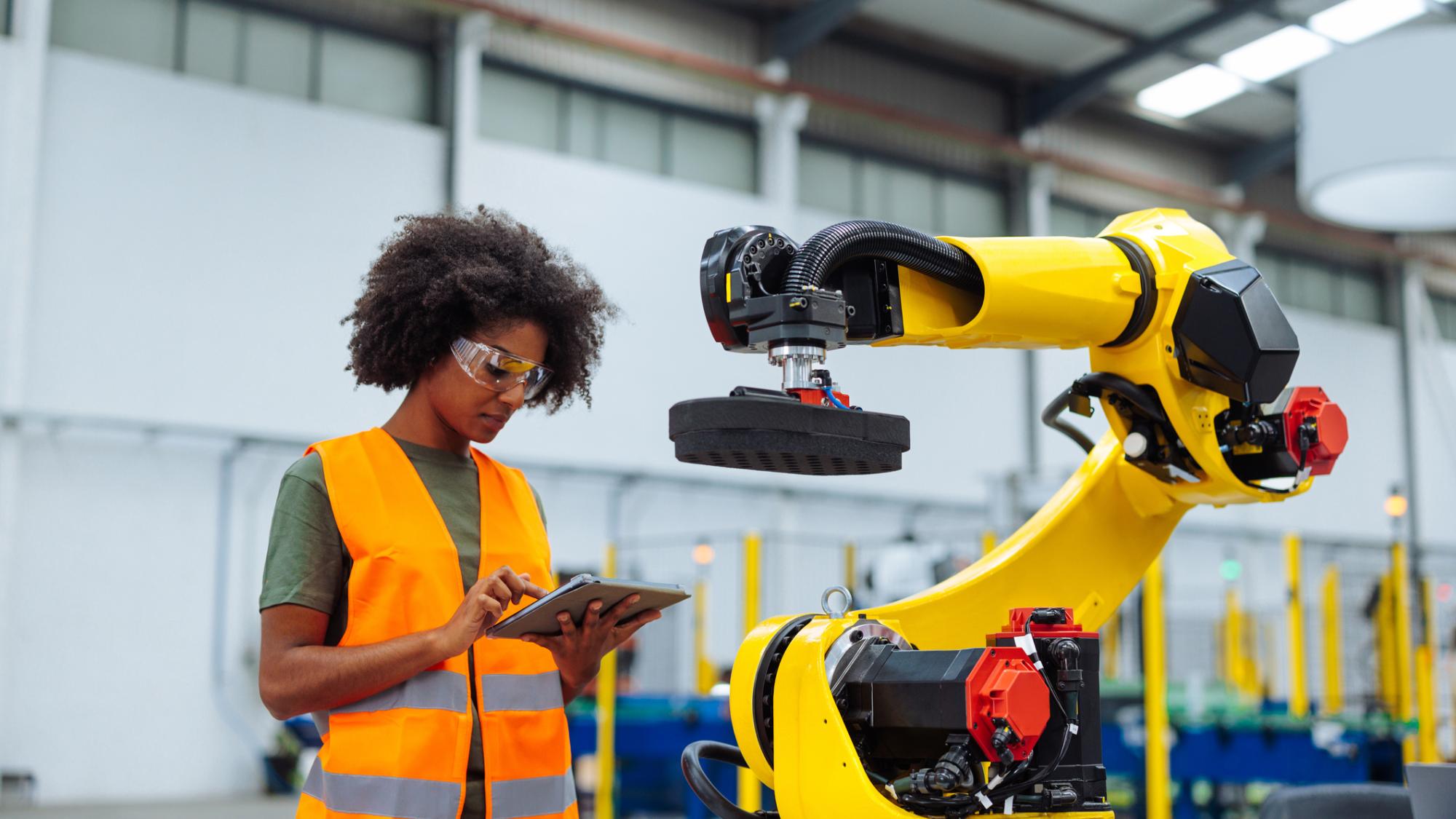 A woman wearing an orange safety vest uses a tablet to program an industrial robotic arm used in manufacturing